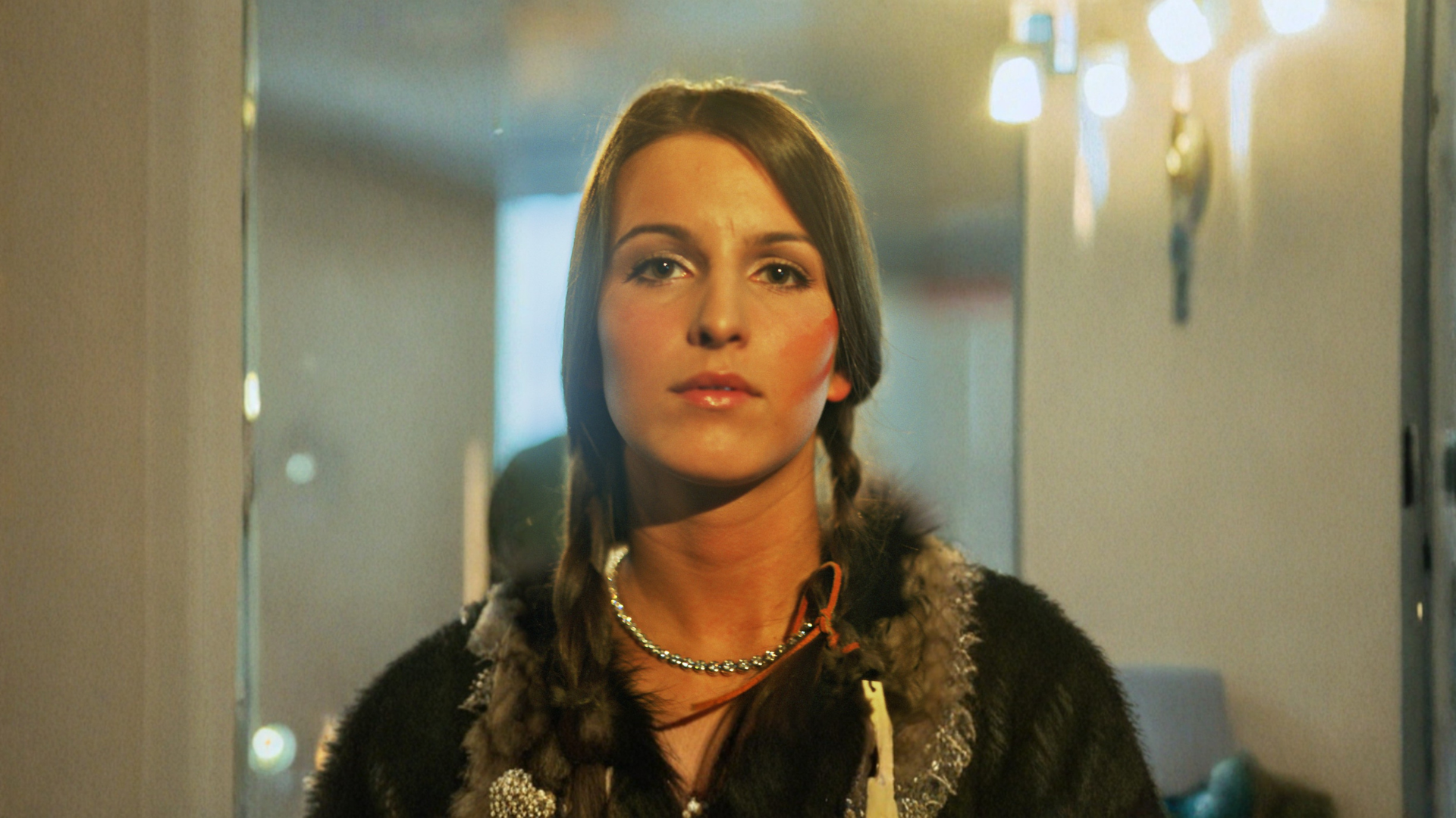 Woman with braided hair wearing a dark coat with fur trim indoors.