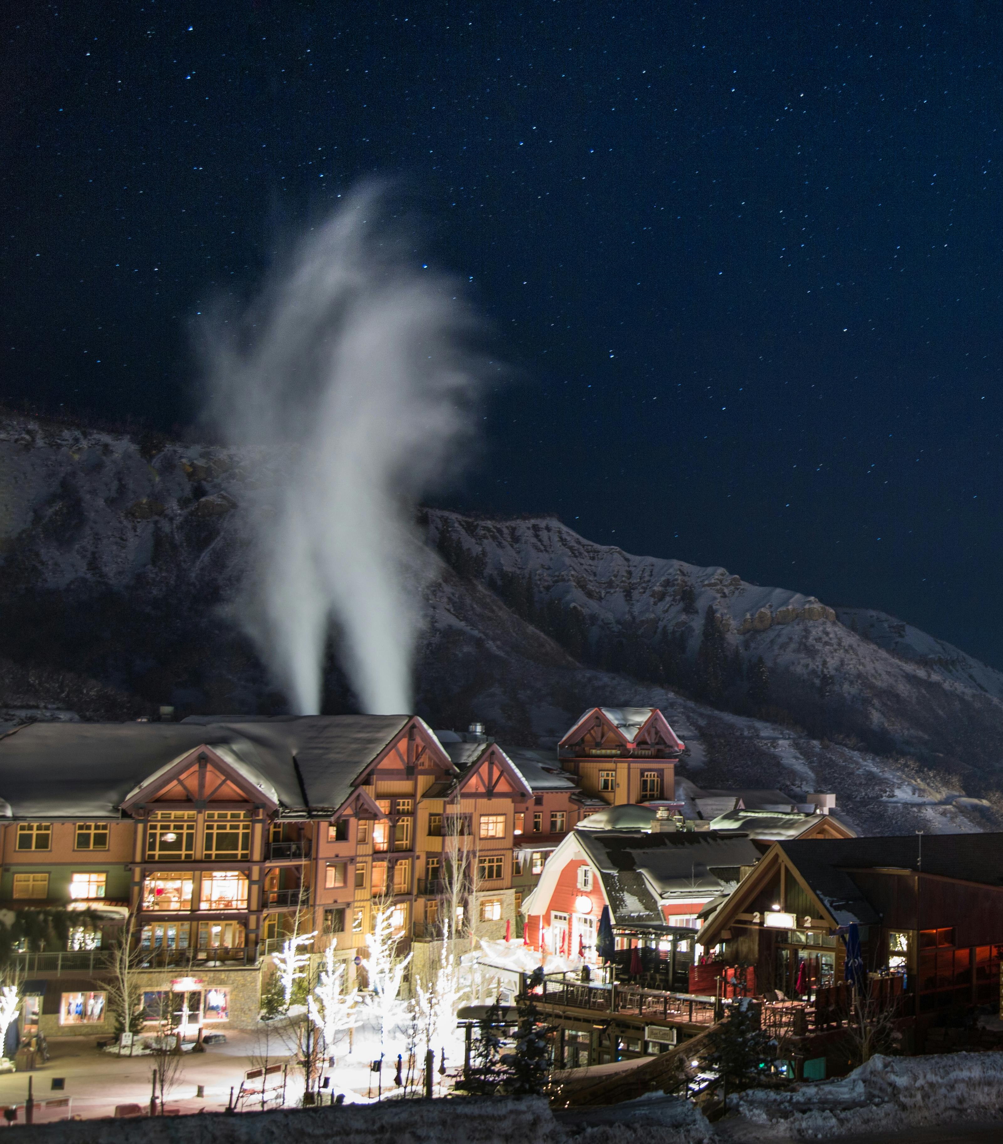 Snowy mountain village at night with illuminated buildings and a large plume of snow being thrown into the air.